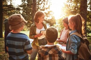 teacher interacting with group of school kids boys and girls during ecology lesson outdoors in forest