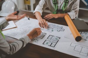 Two architects are discussing house plans in a meeting, pointing at blueprints and diagrams on a wooden table