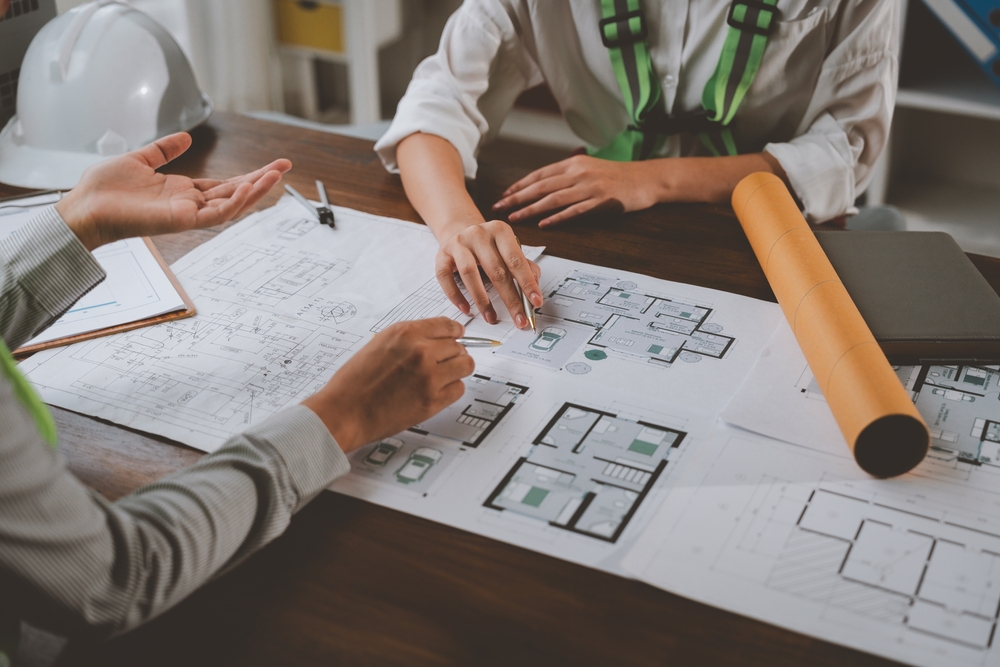 Two architects are discussing house plans in a meeting, pointing at blueprints and diagrams on a wooden table