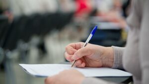 Close-up of adult woman filling out a paper form with a pen while sitting in a classroom or office.