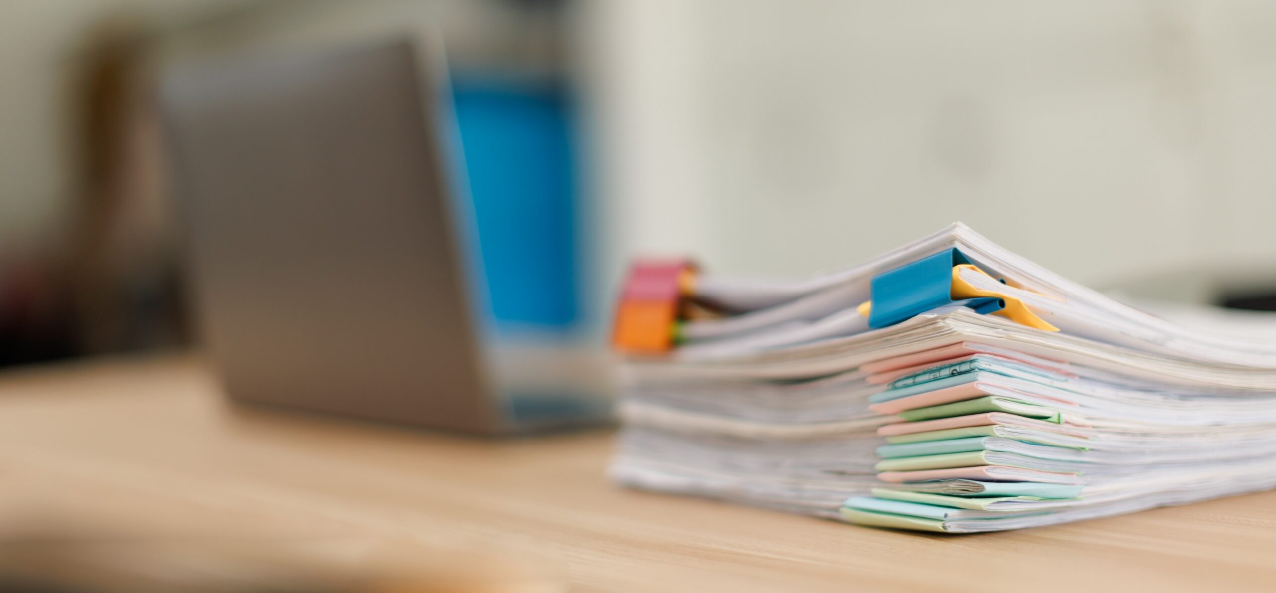 Stack of document files, paperwork, and reports organized with binder clips on a desk next to a blurred laptop, representing business management and data